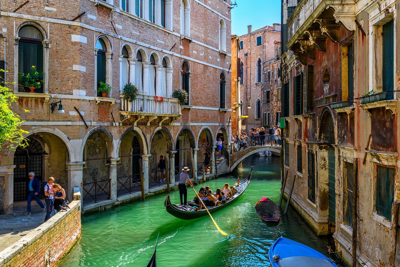 Gondolier steering passengers along a narrow Venetian canal with arched brick buildings and a small bridge.