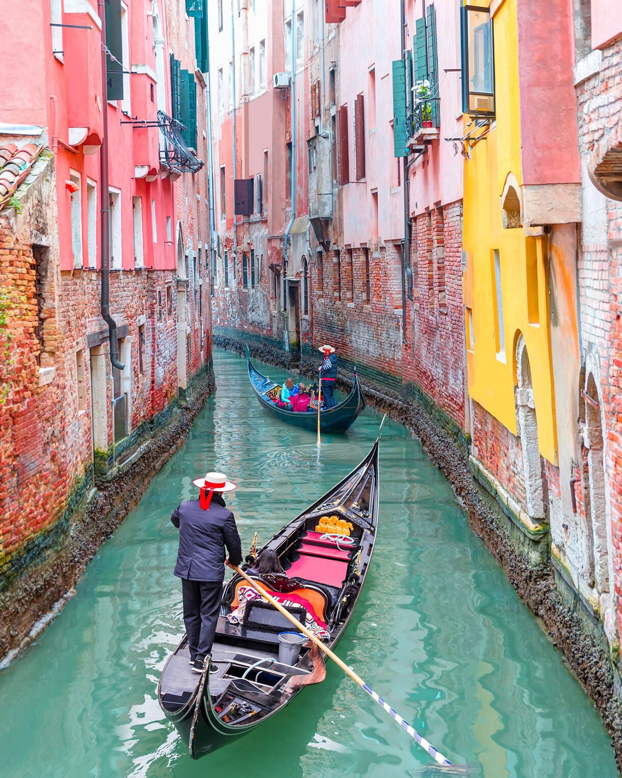 A gondolier in a straw hat steers a black gondola through a narrow Venetian canal lined with colorful brick buildings.
