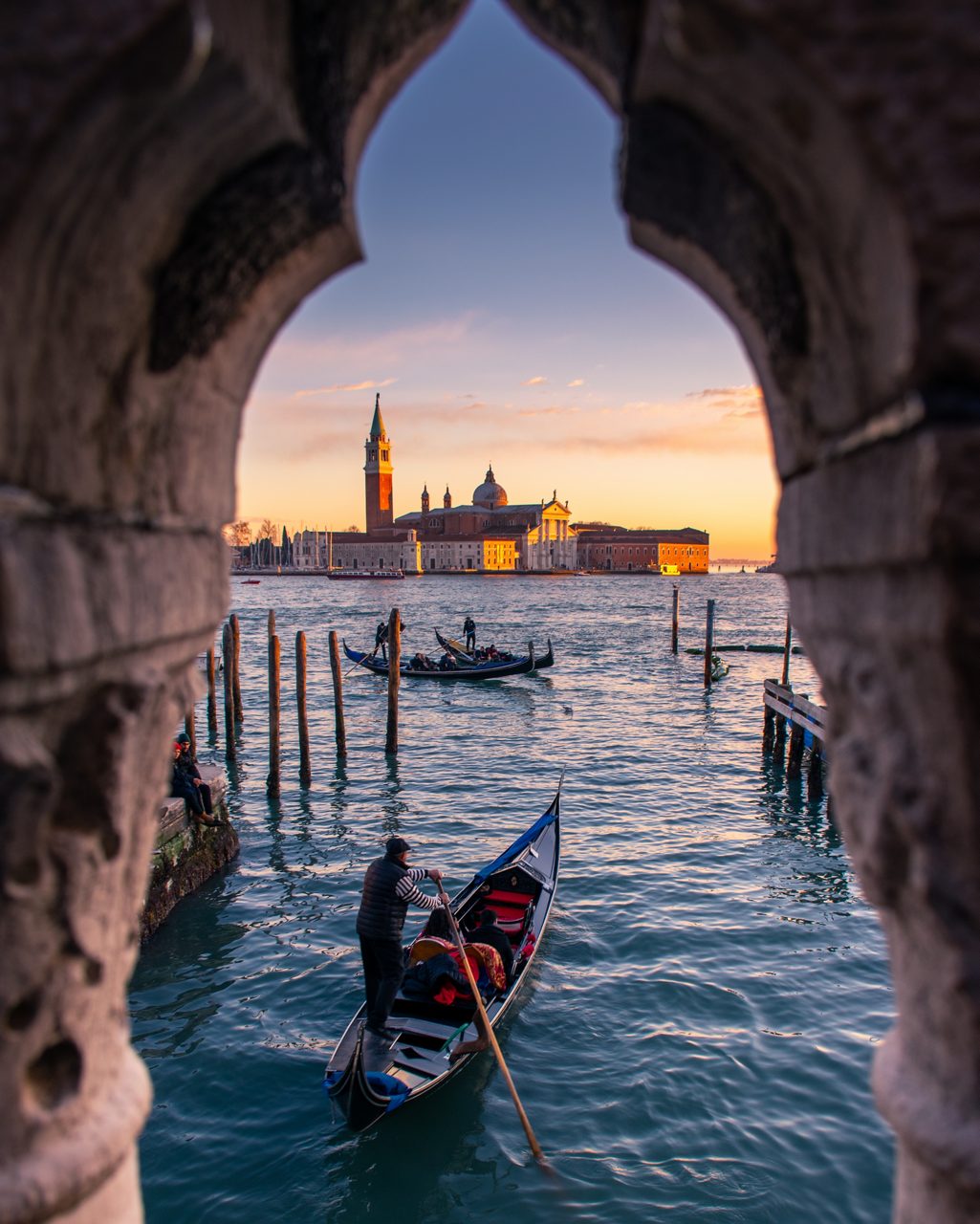 Gondolier rowing at dusk framed by stone archway, with San Giorgio Maggiore lit by the setting sun.