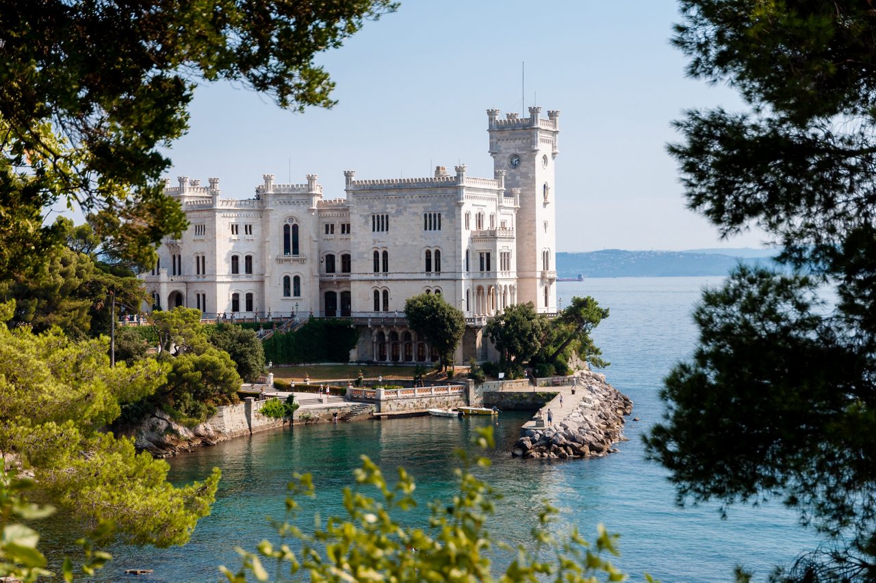 Miramare Castle in Trieste, Italy, a white stone fortress with towers and arched windows perched on a rocky shoreline overlooking turquoise waters.