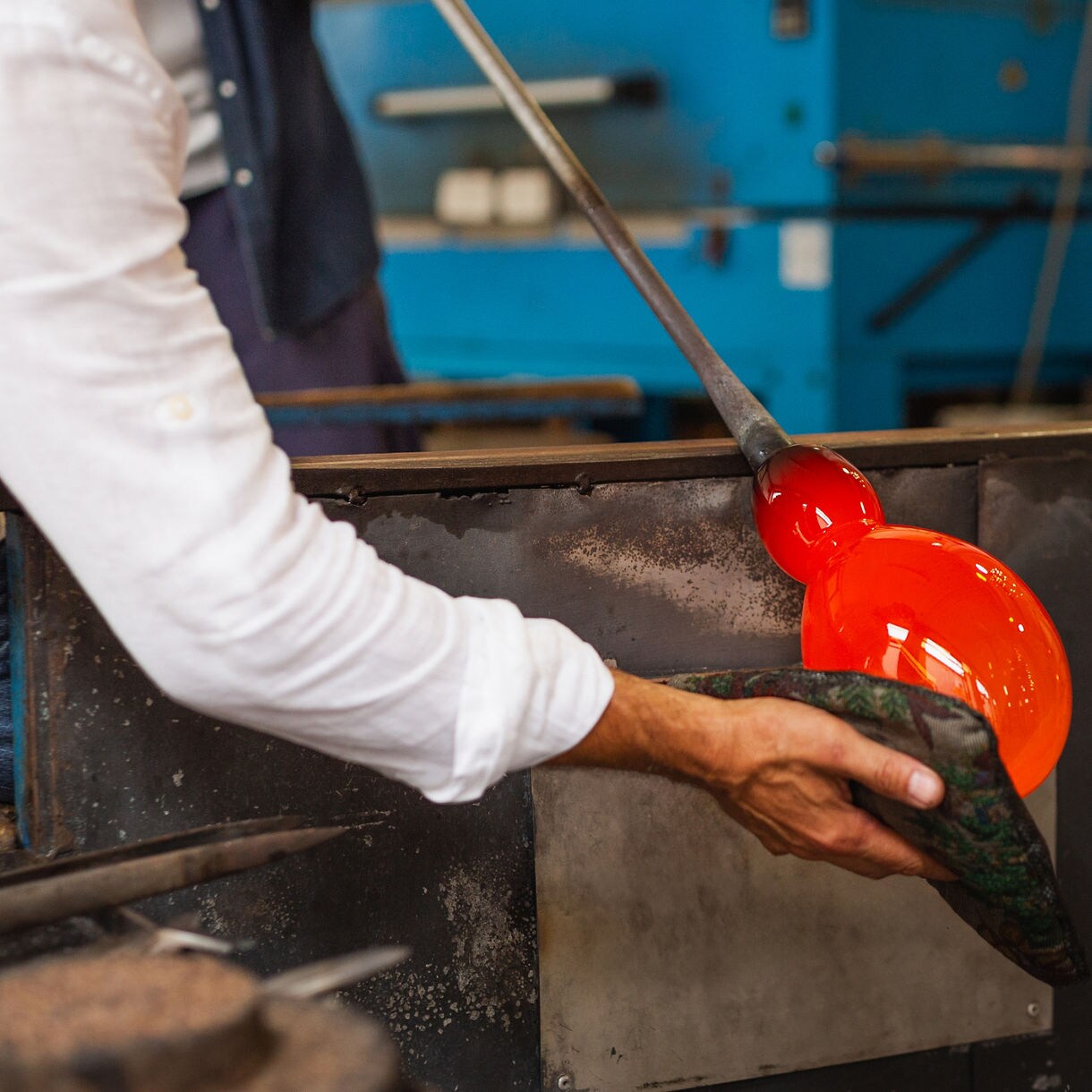 Artisan shaping a glowing ball of molten glass with tools in a Murano workshop.