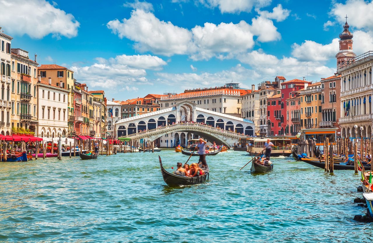 Gondolas and boats on the Grand Canal with the white stone Rialto Bridge and colorful Venetian buildings.