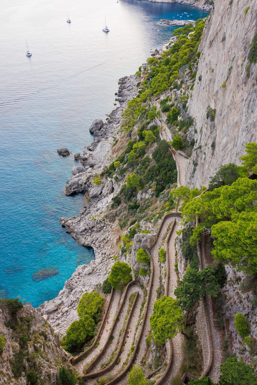 A steep, zigzagging stone pathway along a cliffside on Capri, bordered by green trees above clear blue water and rocky coastline below.
