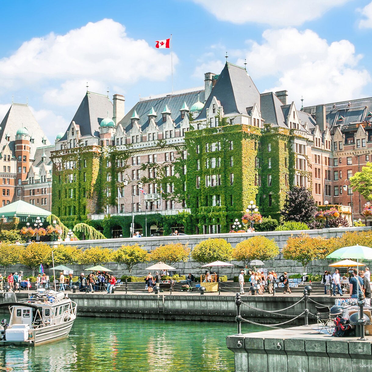 The Fairmont Empress Hotel in Victoria, British Columbia, draped in ivy, with a clear blue sky above and a bustling waterfront filled with people and boats in the foreground.