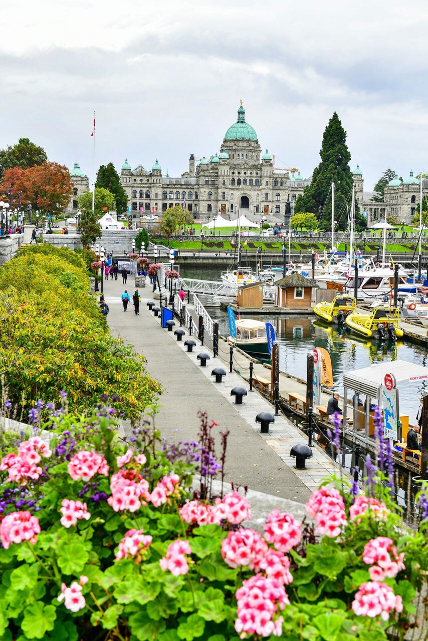 A scenic view of Victoria’s Inner Harbour, with colorful flowers in the foreground, a bustling marina and the grand British Columbia Legislature building in the background under a cloudy sky.