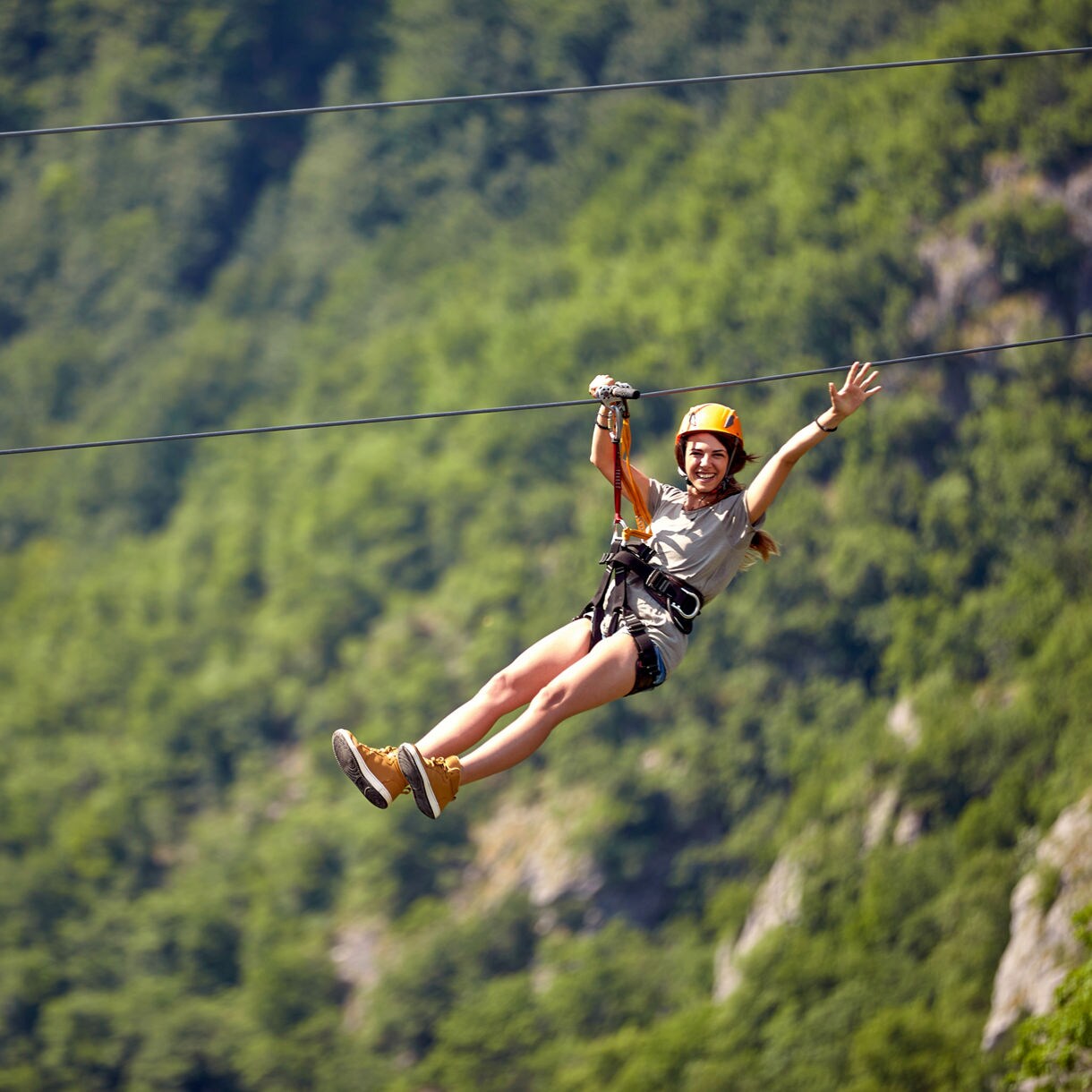 A smiling woman zipping across a lush forested landscape on a zipline, wearing a helmet and harness with green hills and trees in the background.