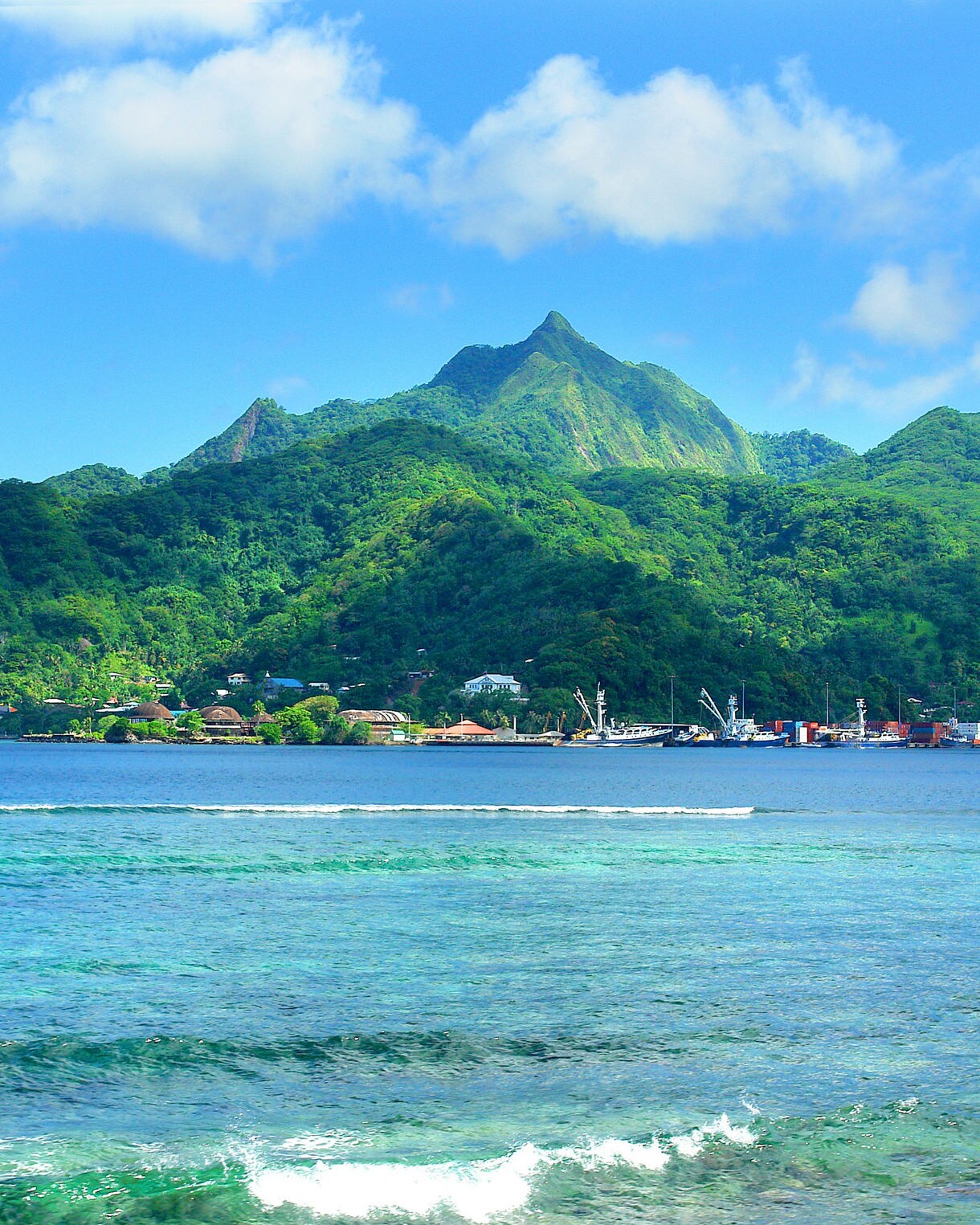 A clear view of a lush, green mountain range behind a calm blue bay with gentle waves and a small coastal town along the shoreline.