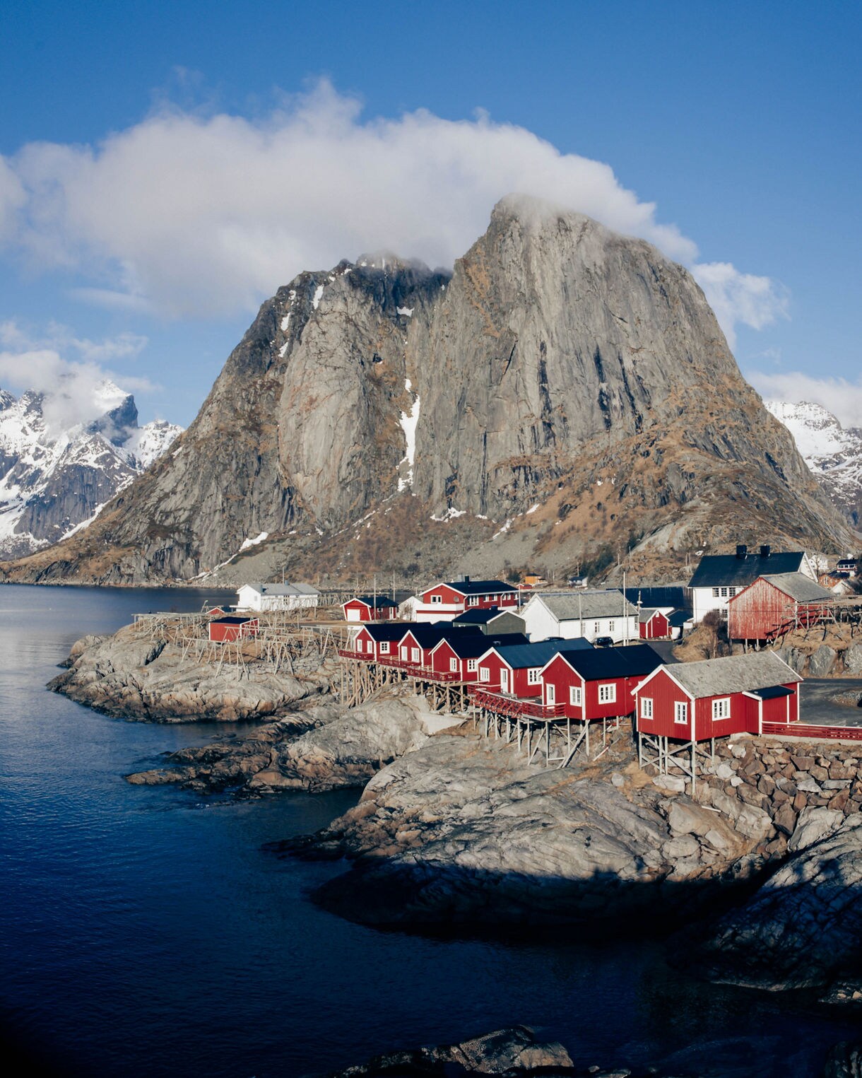 A cluster of red wooden cabins on stilts sits along a rocky shoreline beneath a massive mountain in Norway’s Lofoten Islands.