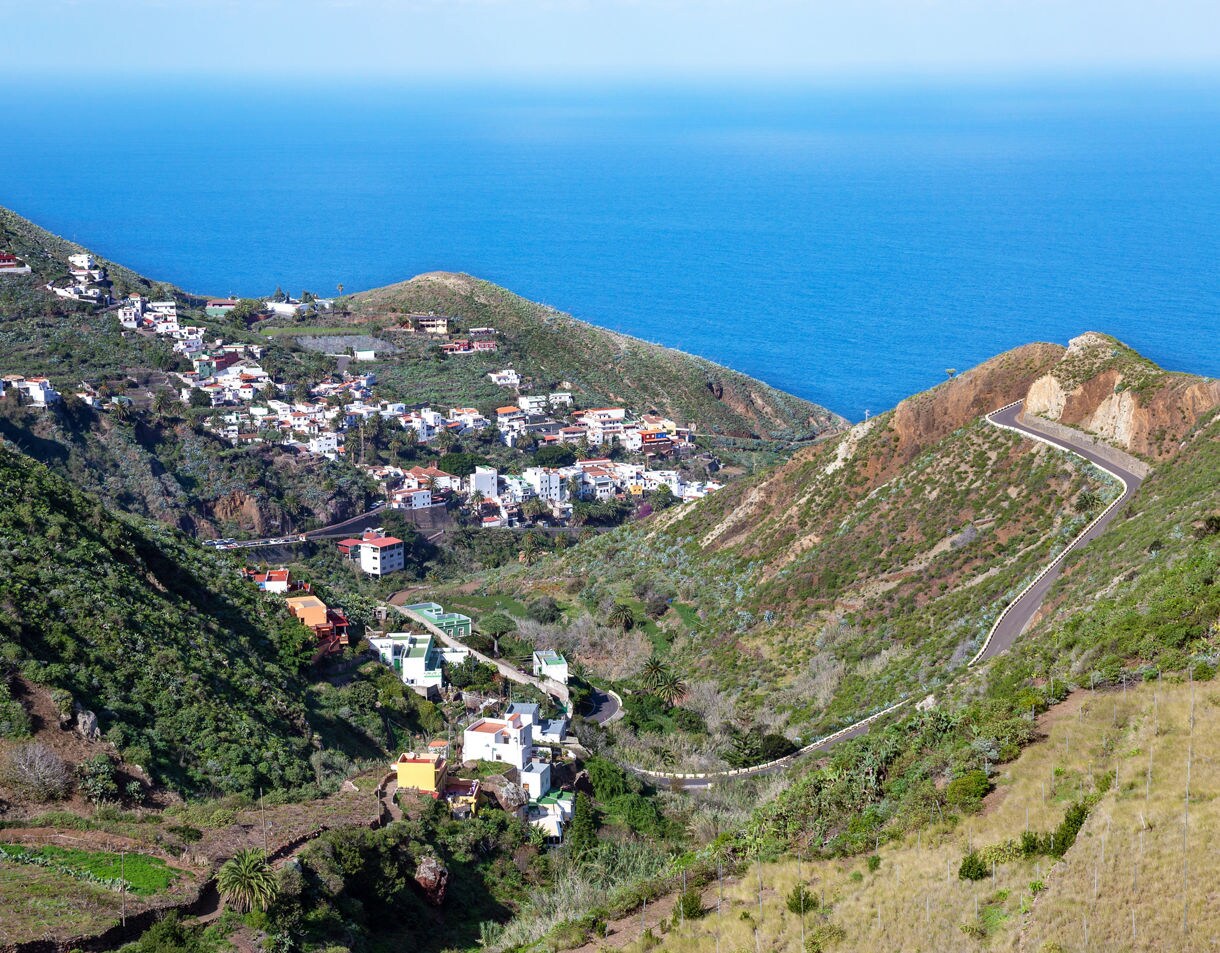 Aerial view of Taganana village in Tenerife with white houses, green hills and a winding road leading toward the blue ocean.