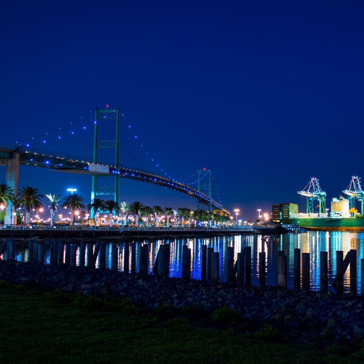 Night view of Vincent Thomas Bridge and the Port of Los Angeles, with colorful lights reflecting on the water.