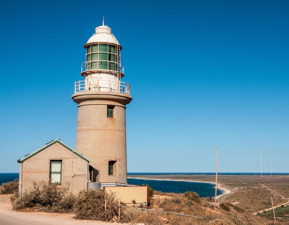 A weathered lighthouse and small building overlooking a sweeping coastal landscape with blue ocean and sandy shoreline under a clear sky.