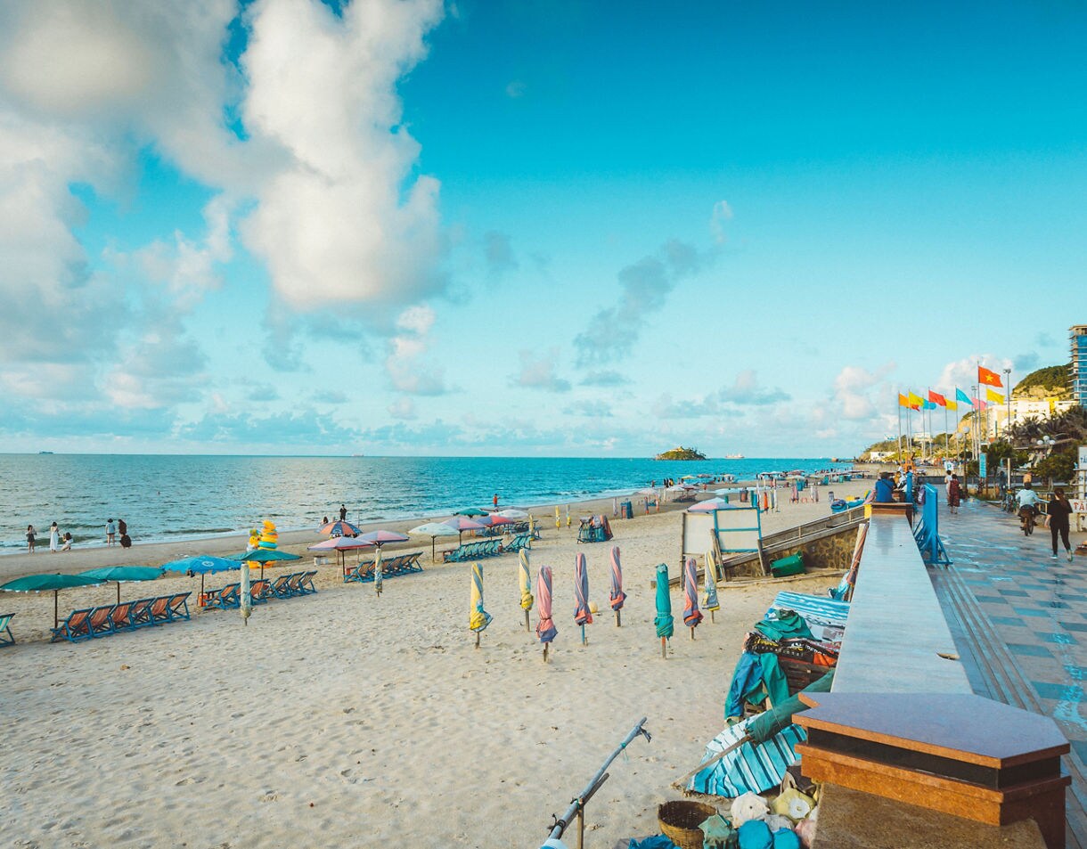 Colorful umbrellas and lounge chairs line the sandy shore of Vũng Tàu Beach as visitors stroll by the calm blue sea.