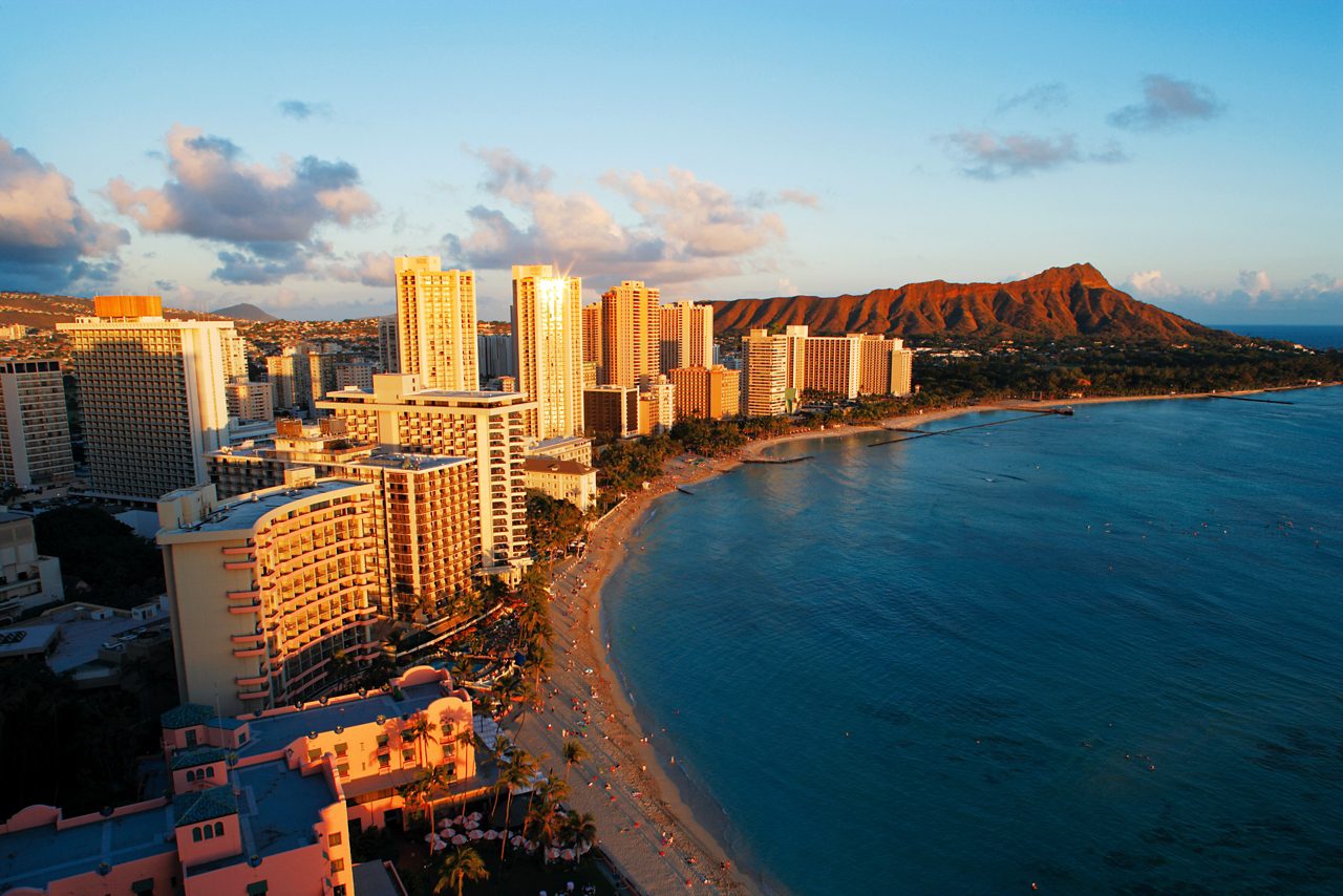 Aerial view of Waikiki’s beachfront skyline in Honolulu, Hawaii, at sunset, with hotels and buildings illuminated by the golden light, and the famous Diamond Head volcanic crater in the distance along the coastline.