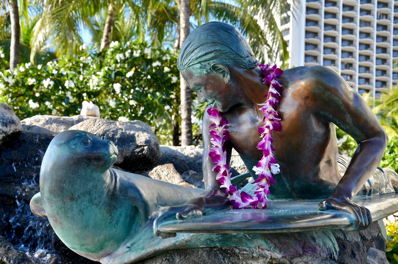 Bronze statue of a surfer with a lei around his neck leaning toward a Hawaiian monk seal at Waikiki, surrounded by palm trees.