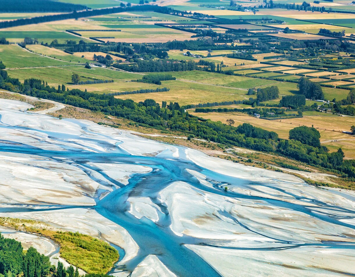 Aerial view of the Waimakariri River’s intertwining blue streams flowing across pale sandbanks with farmland in the background.