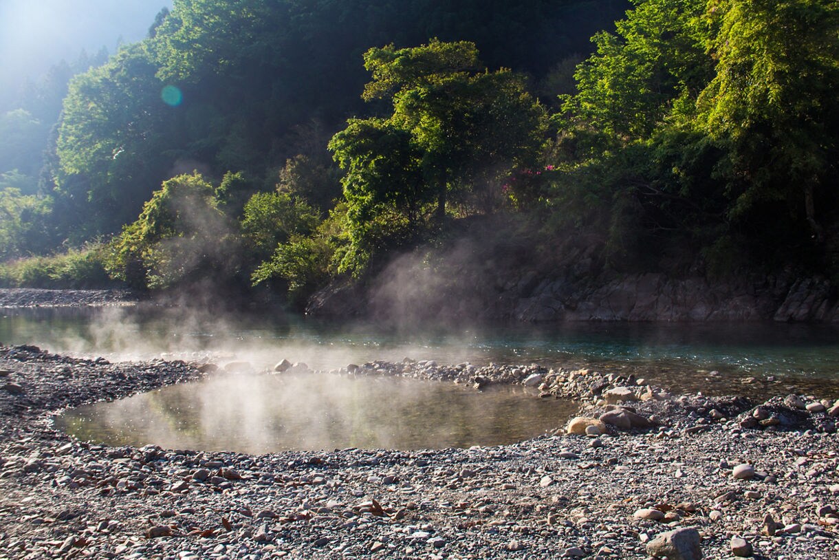Steaming natural hot spring pool beside a rocky riverbank surrounded by dense green forest.