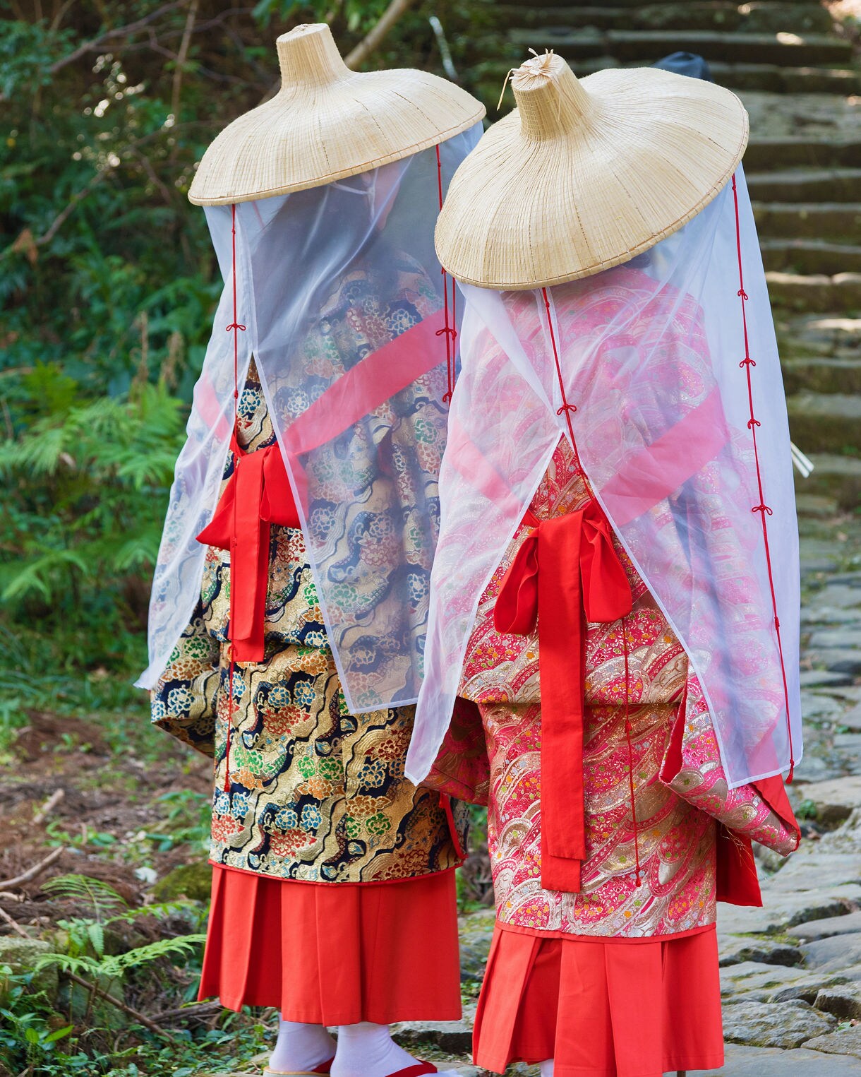 Two people wearing traditional Japanese religious or ceremonial costumes with large woven conical hats, white translucent veils, and colorful layered robes in red and patterned fabrics, standing on a stone path in a garden setting with traditional architecture visible in the background.