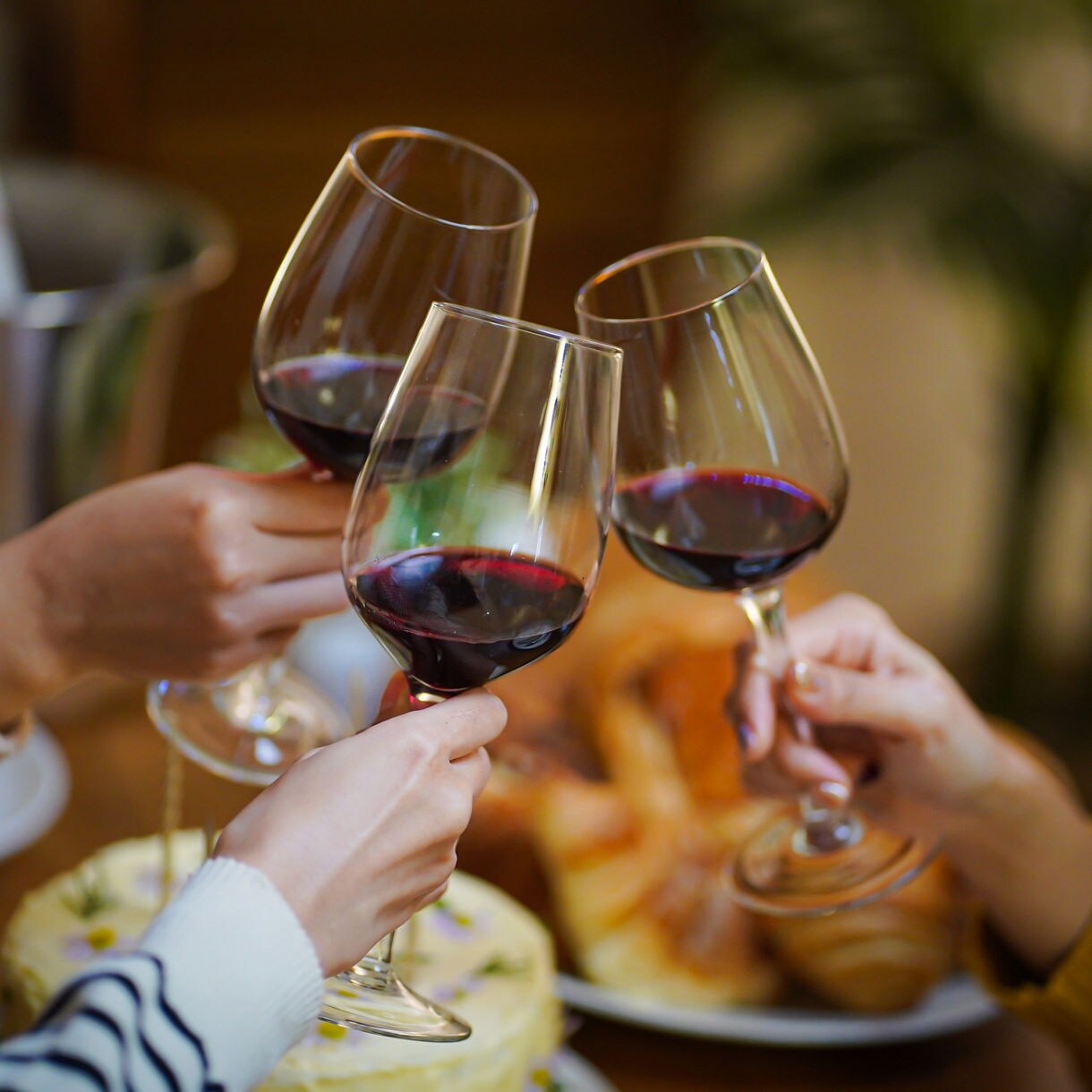 Three people clinking glasses of wine over a table with food and dessert in a cozy setting.