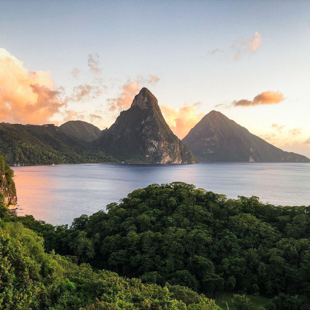 Twin volcanic peaks known as the Pitons rise above calm blue water at sunset, surrounded by lush rainforest and soft orange clouds in St. Lucia.