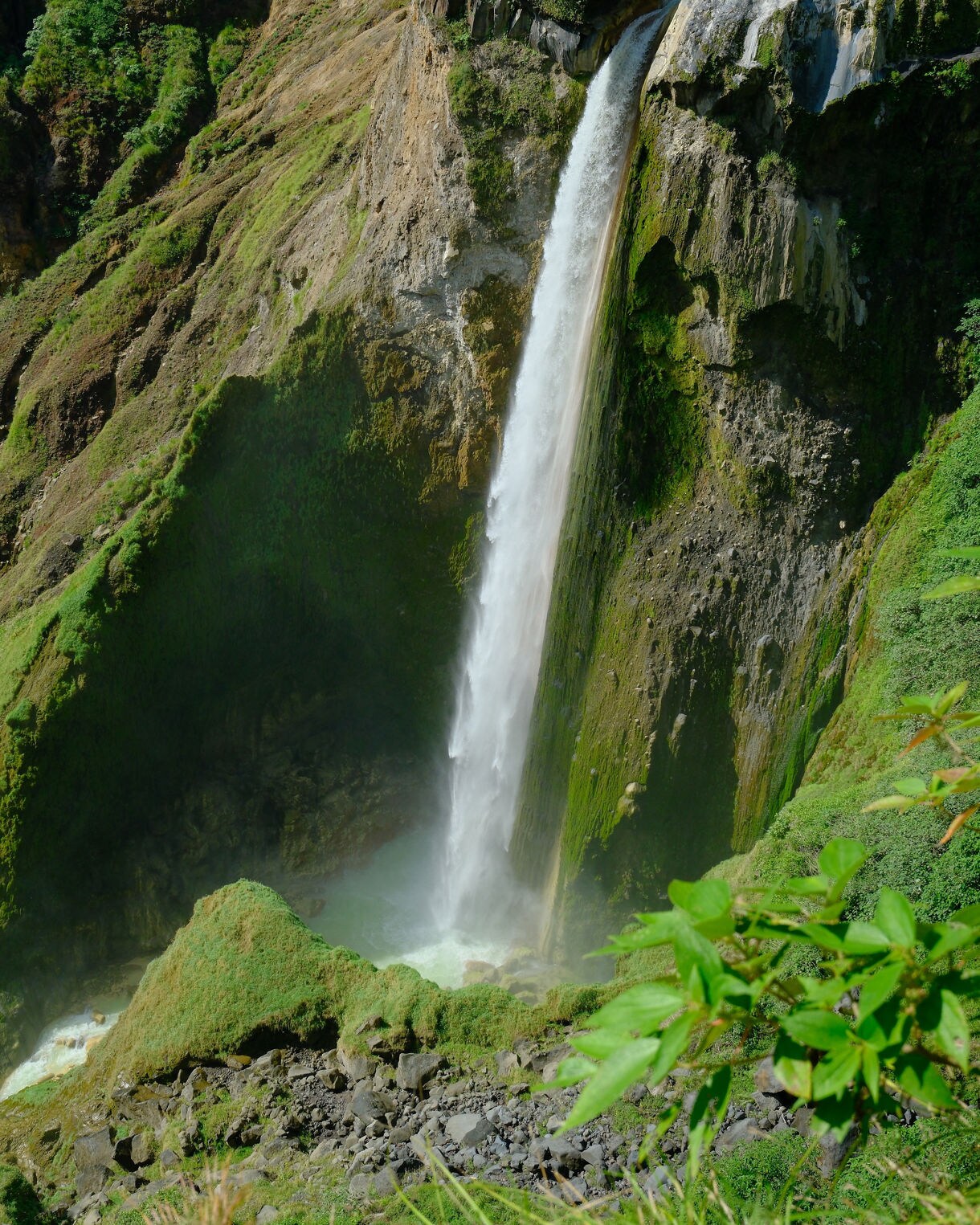 Tall waterfall plunging down a steep green cliff into a rocky turquoise pool, surrounded by moss-covered walls and dense tropical vegetation.