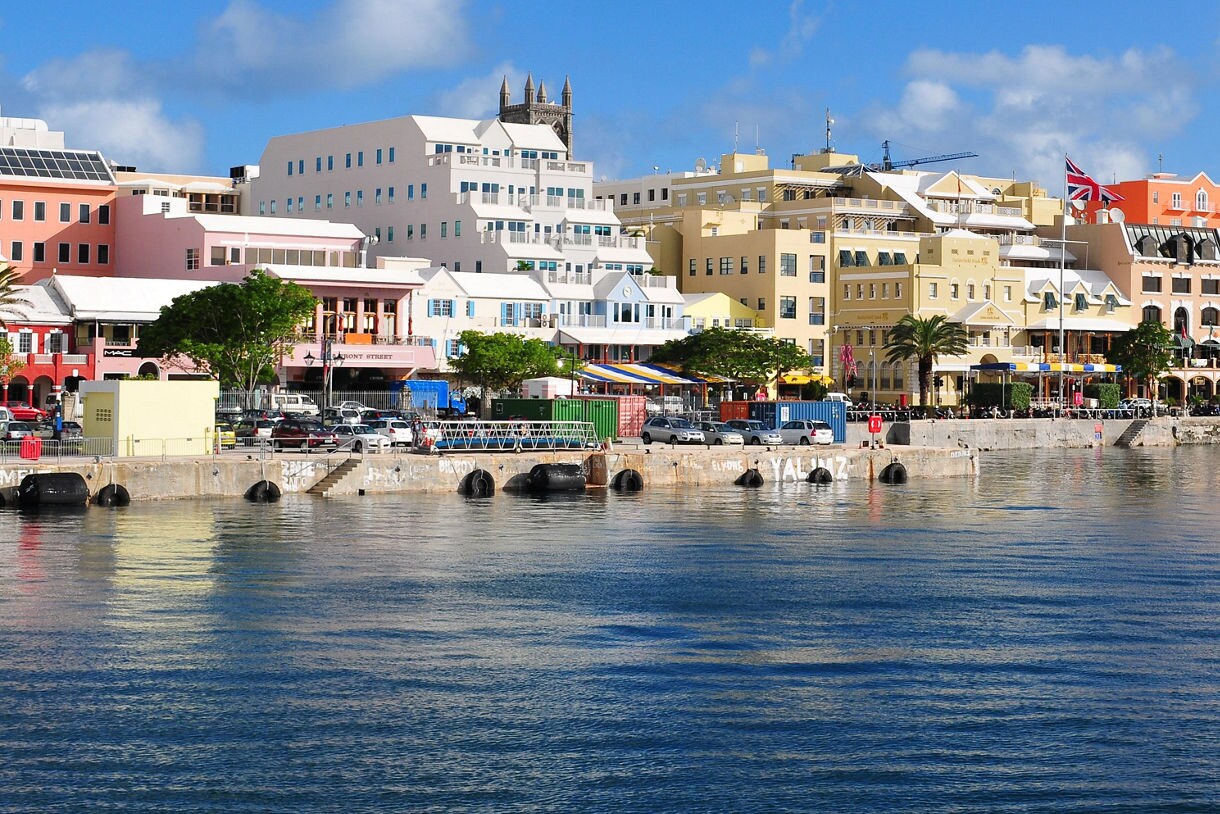 Waterfront view of Hamilton, Bermuda with rows of pastel buildings, white roofs and the Union Jack flag overlooking the harbor.
