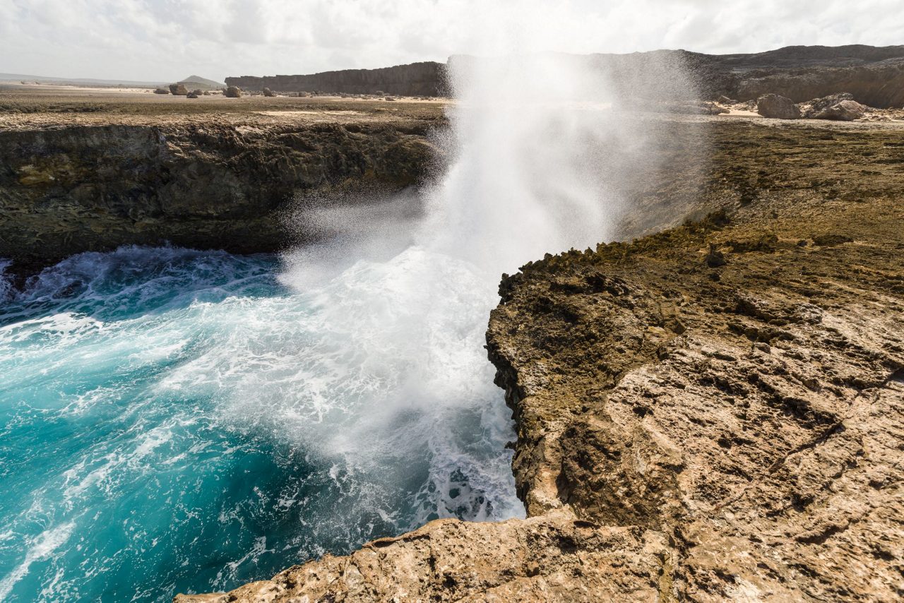 Dramatic coastal scene in Bonaire showing powerful waves crashing against rugged limestone cliffs with spray shooting high into the air under a cloudy sky.