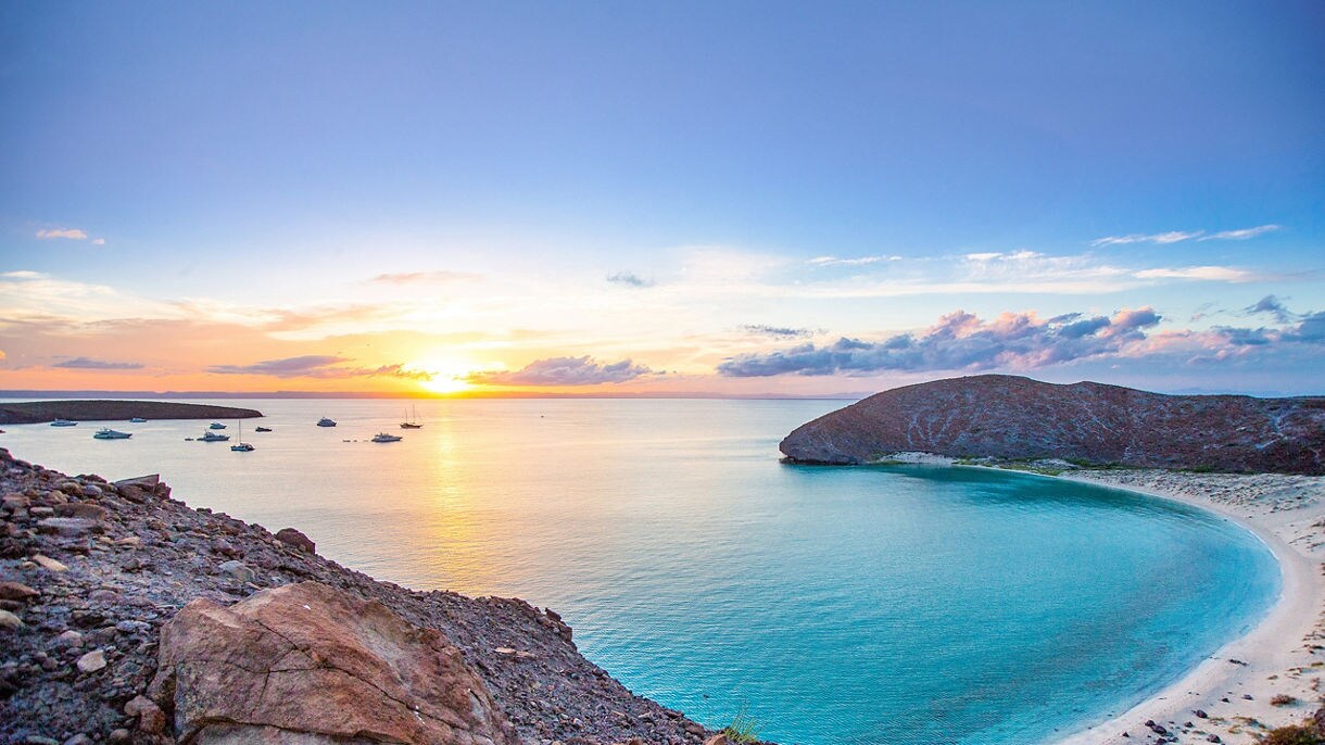 A curved Mexican beach cove with turquoise waters surrounded by rocky cliffs at sunset, with several boats anchored in the distance.