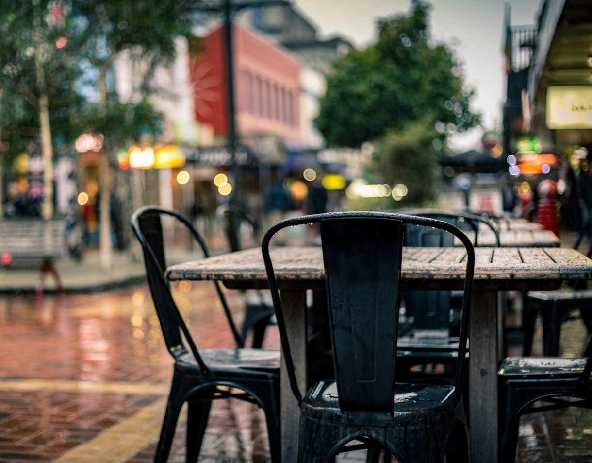 Close-up of a wet outdoor café table and chairs on a rainy day in Wellington, with blurred city lights and pedestrians in the background.