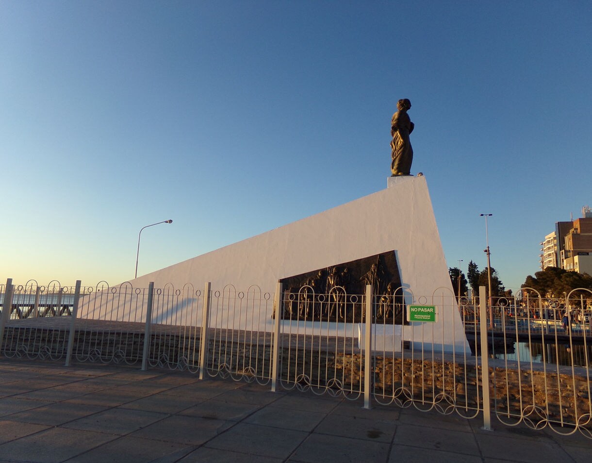 A waterfront monument in Puerto Madryn featuring a tall white structure topped with a bronze statue, lit by warm late-day sunlight.