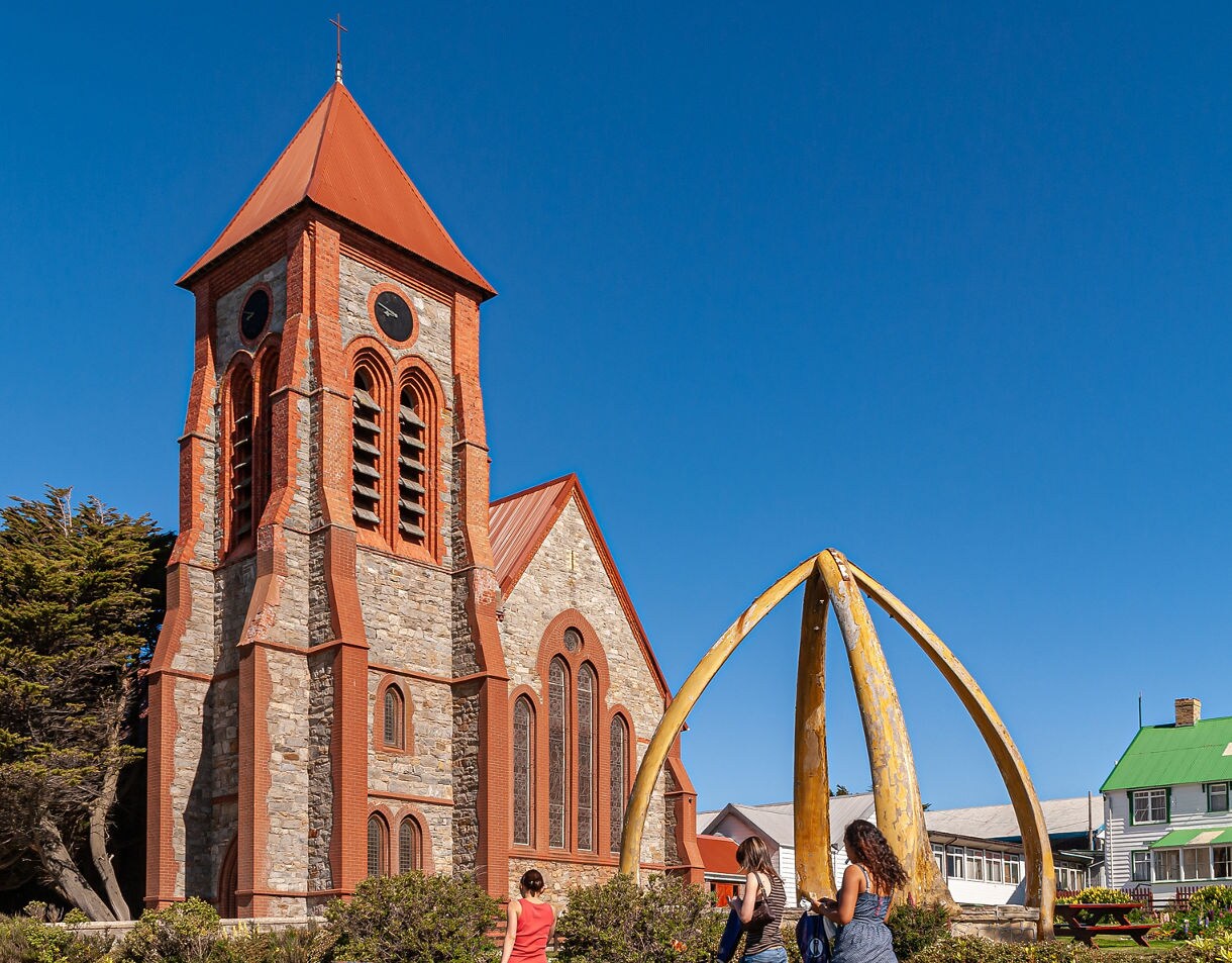 Stone Christ Church Cathedral with a tall red-roofed tower beside a large yellow whalebone arch, with three people walking in the foreground under a clear blue sky.