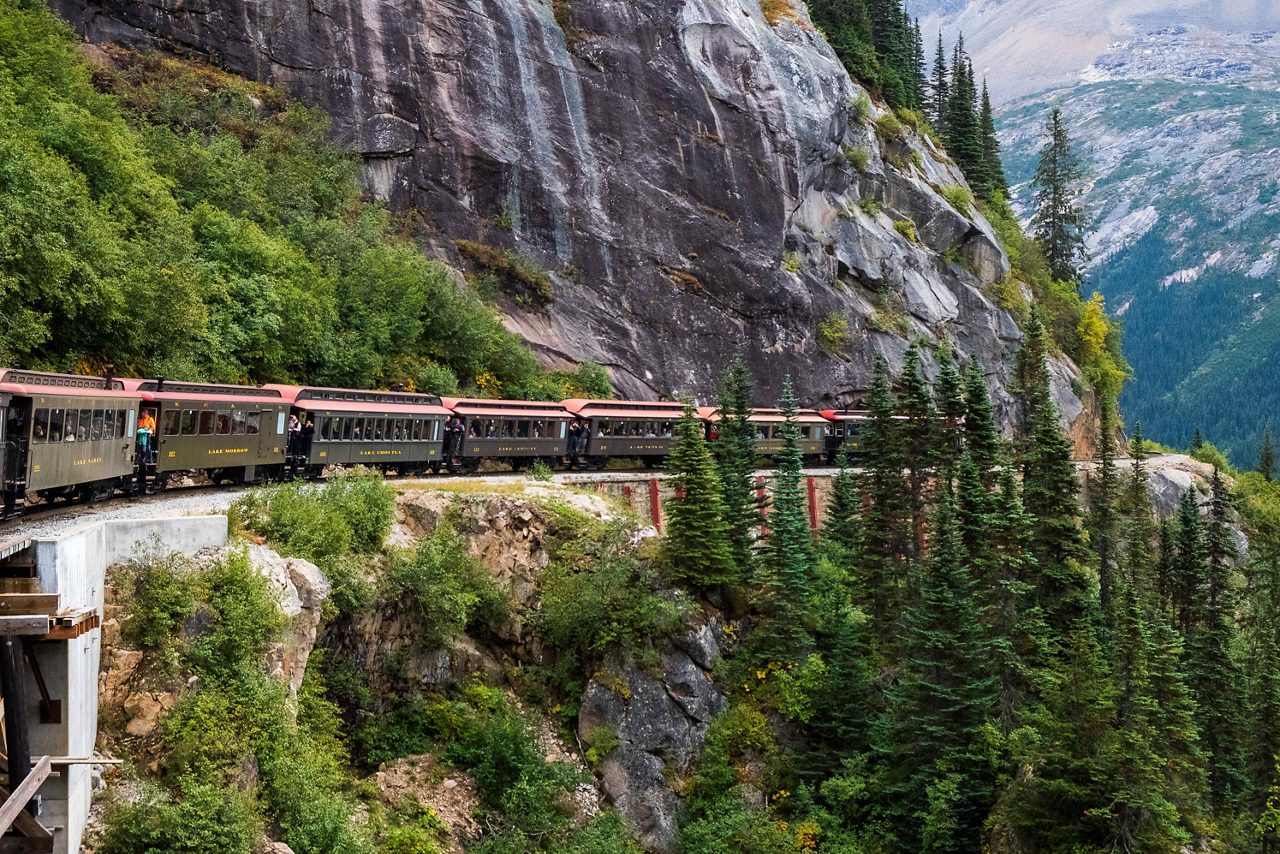 Vintage train traveling along a steep mountain pass with pine trees, rocky cliffs and distant alpine scenery.