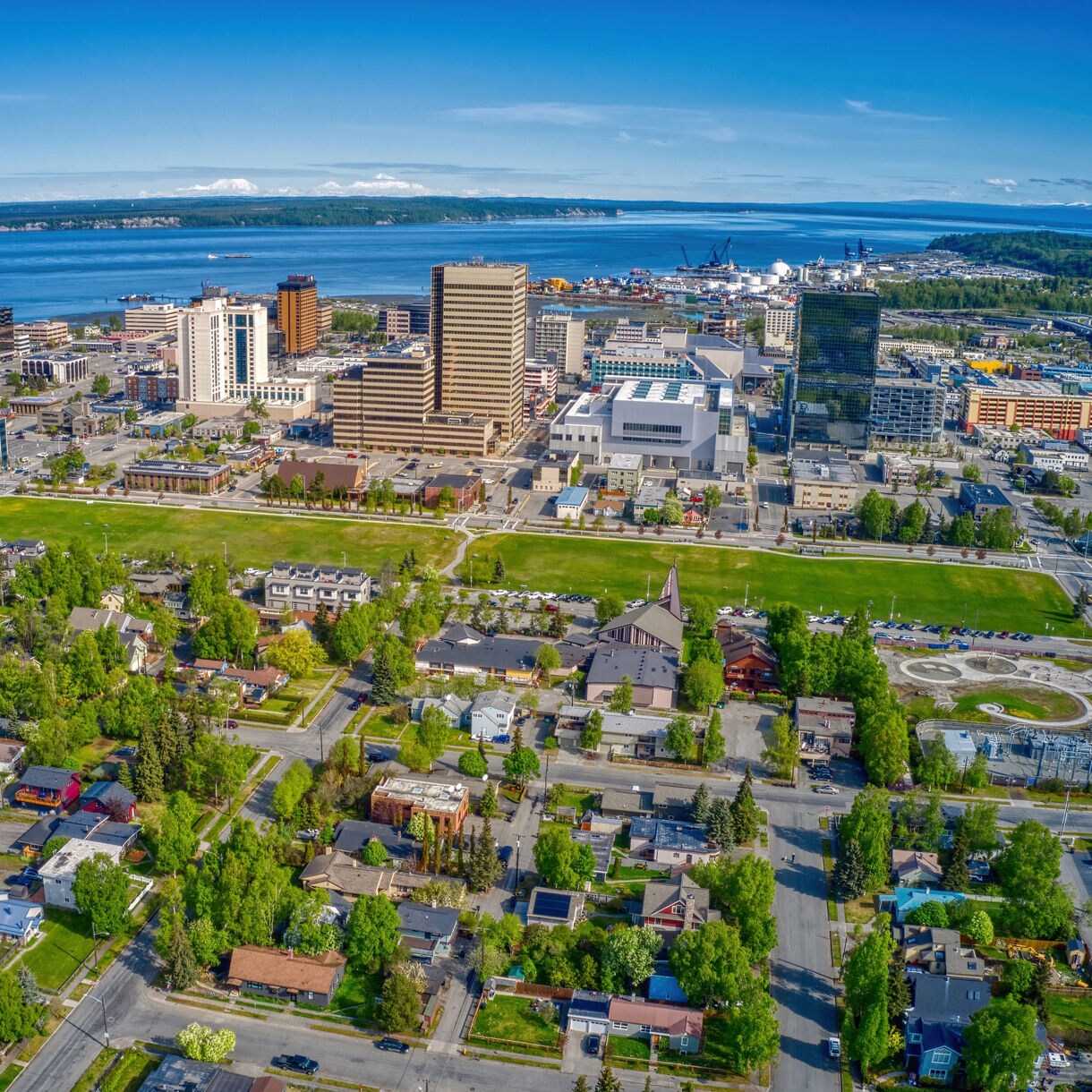 Aerial view of downtown Anchorage, Alaska on a clear summer day, showing modern buildings, green residential neighborhoods, and the blue waters of Cook Inlet in the background.