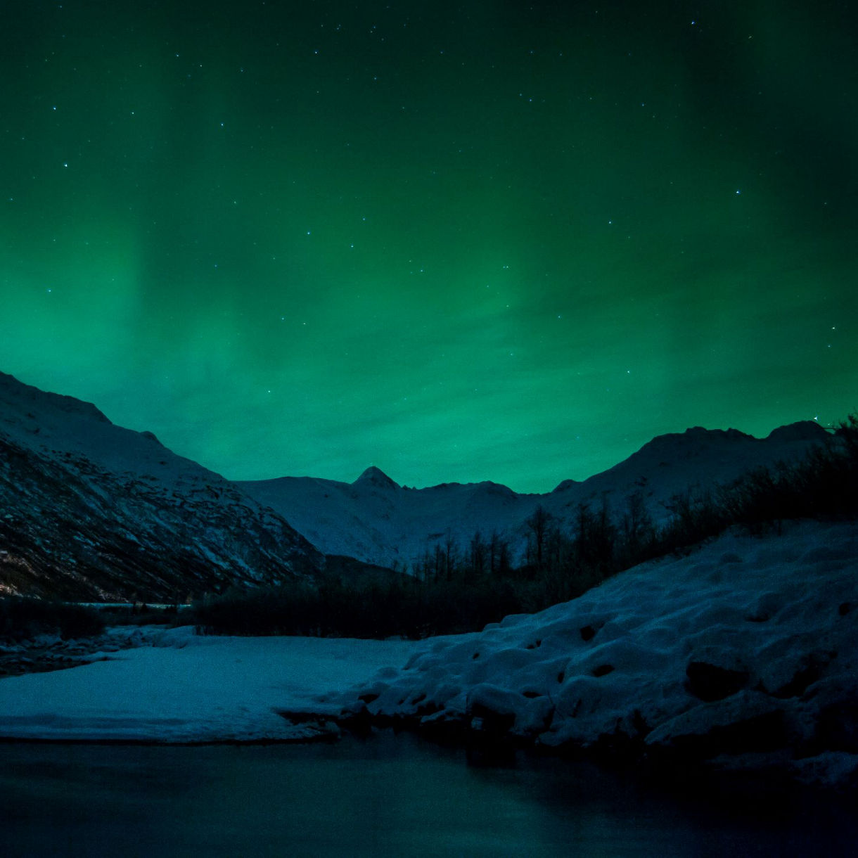 Green northern lights illuminate a dark winter landscape with snow-covered mountains and a partially frozen stream.