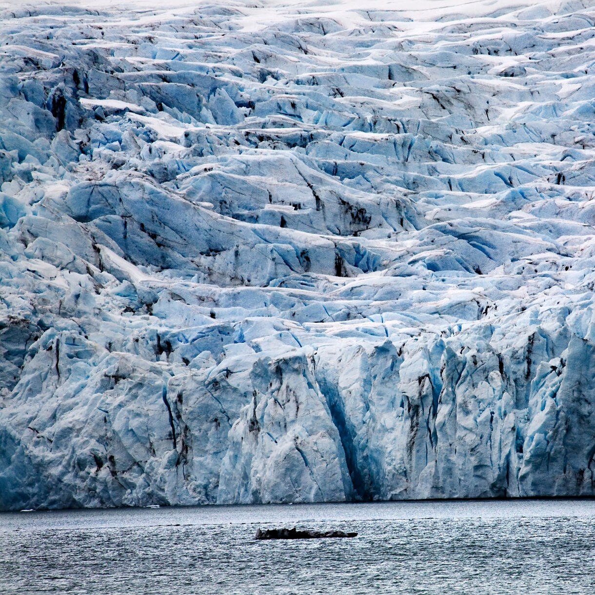 Massive blue and white ice formations of Portage Glacier rising above the water, with deep crevasses and dark striations visible in the frozen surface.