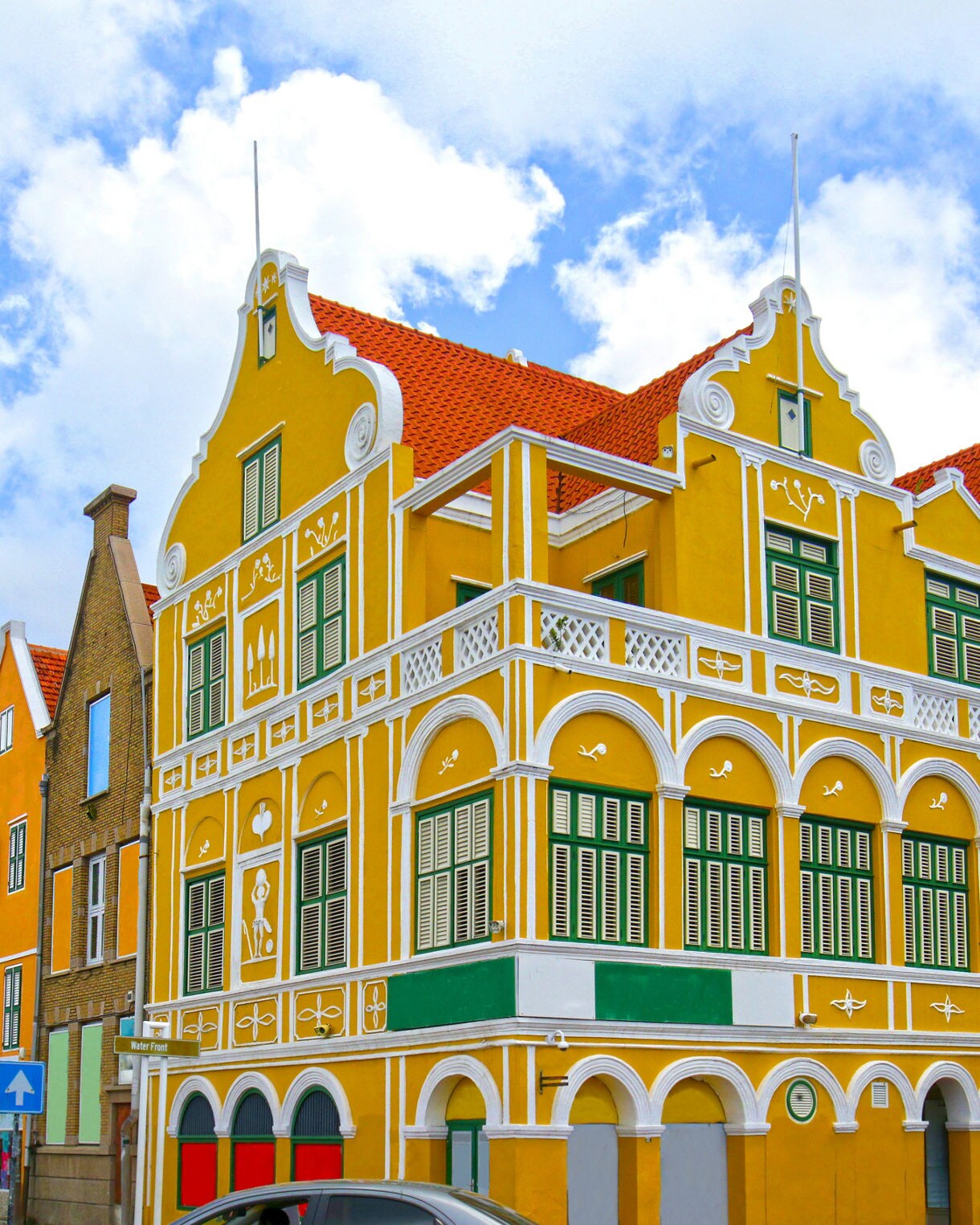 A row of brightly colored Dutch colonial-style buildings in Willemstad, Curaçao, featuring striking yellow, green, and pastel facades with ornate architectural details and red-tiled roofs.