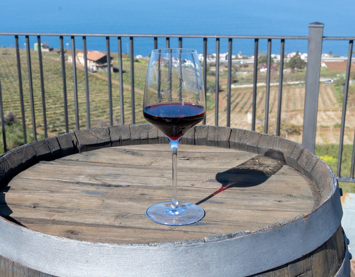 Glass of red wine on a wooden barrel with terraced vineyards and the Atlantic Ocean in the background on a sunny day in Tenerife.