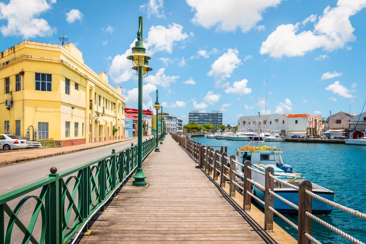Wooden pier with green metal railings extending over blue harbor water with moored boats, yellow colonial building on the left, and various waterfront structures under a cloudy blue sky.