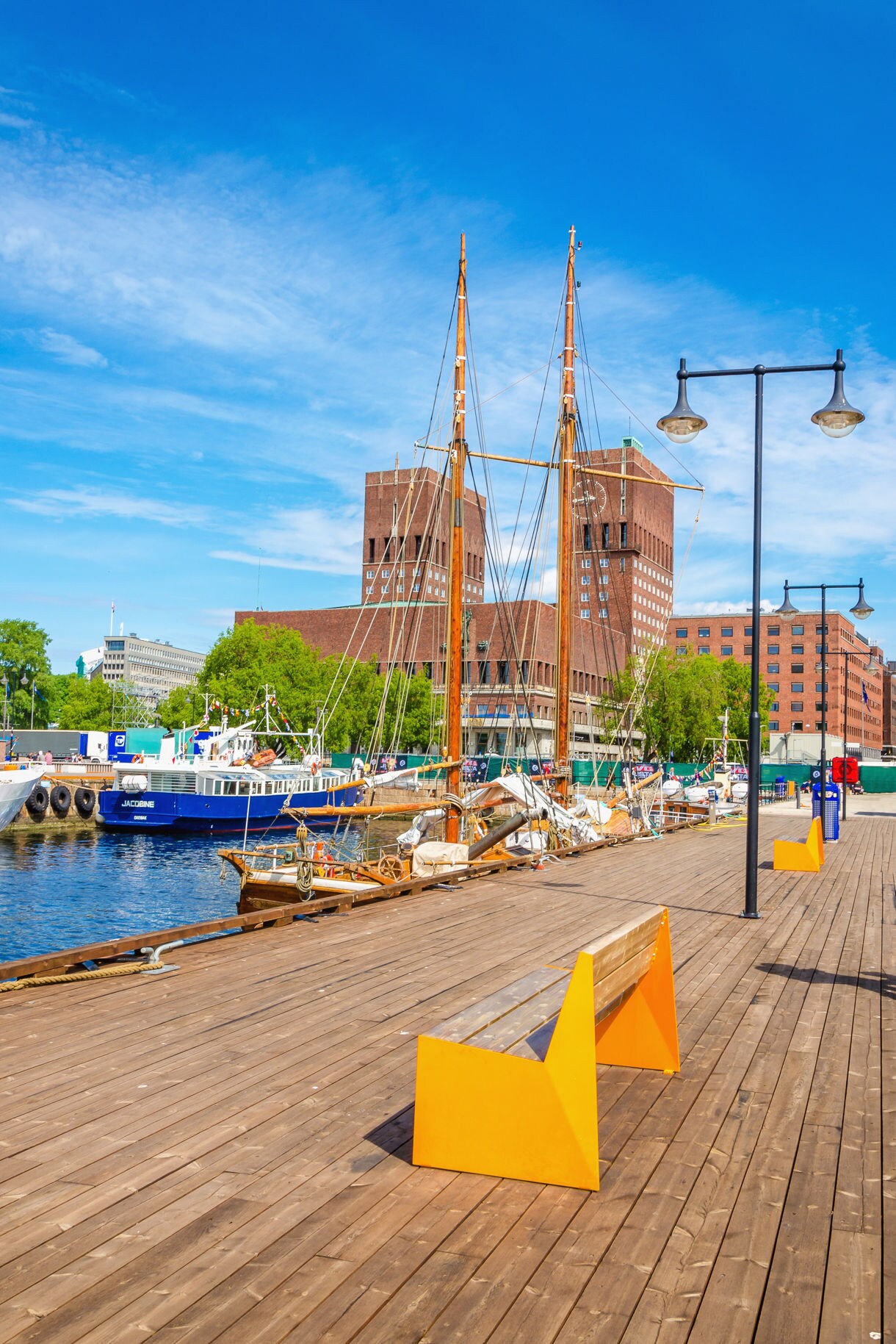 Wooden pier along Oslo’s harbor with moored sailboats and bright benches, facing the twin-towered City Hall under a clear blue sky.