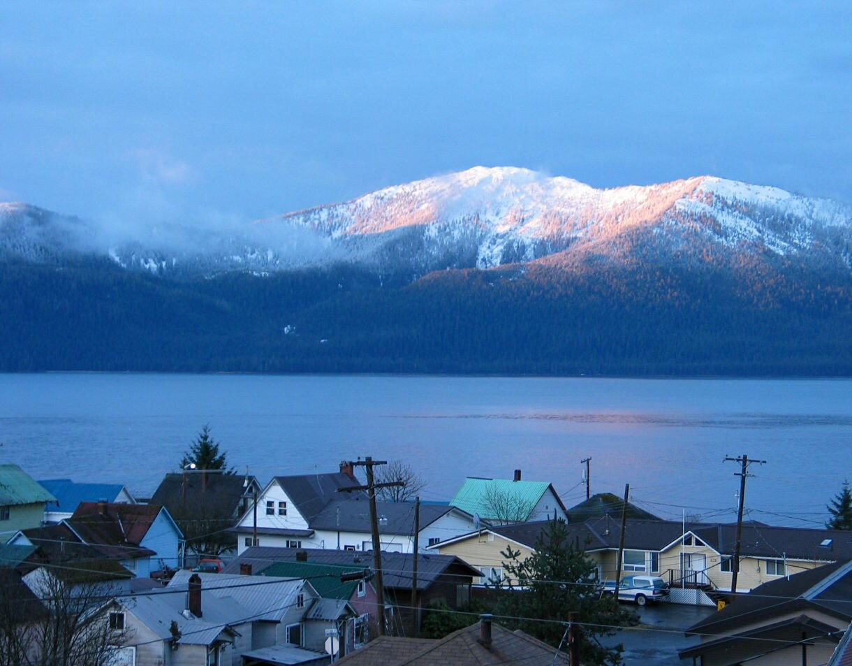 Aerial view of Wrangell, Alaska showing a small residential community with colorful houses and green metal roofs situated along a large body of water, with dramatic snow-covered mountains rising in the background under a clear blue sky during sunset lighting.