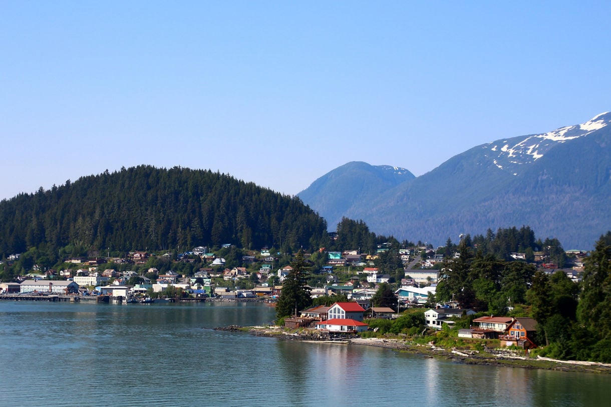 A picturesque view of Wrangell, Alaska, featuring colorful homes and historic buildings along the waterfront, surrounded by dense forests and towering mountains under a clear blue sky.