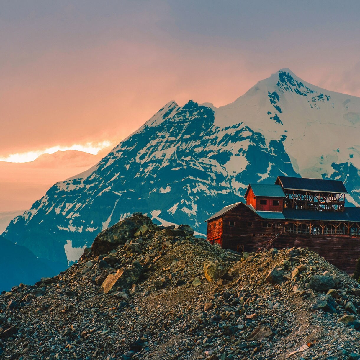 Abandoned wooden mining structure perched on a rocky ridge in Wrangell Alaska, with snowcapped mountains, evergreen forest and a glowing sunset sky in the background.