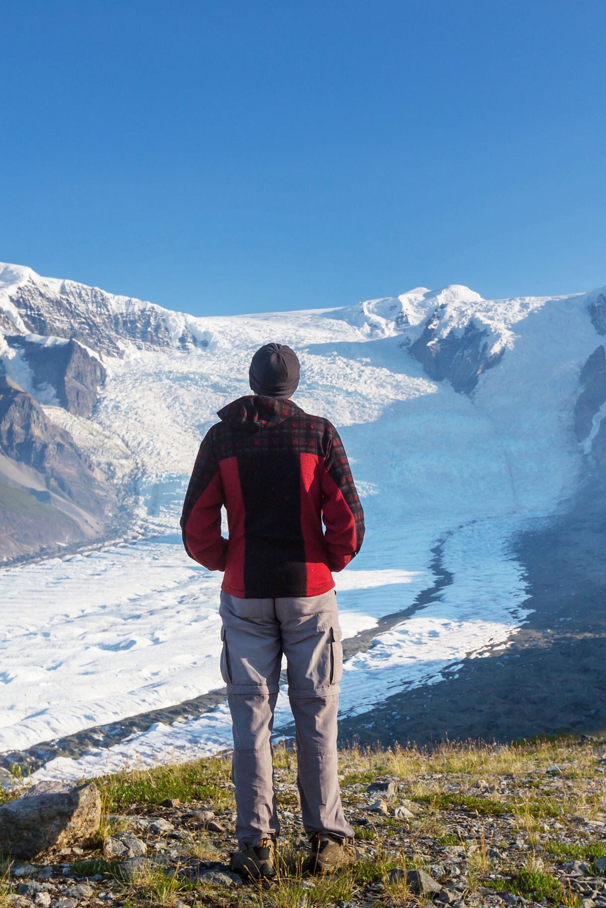 Person in red jacket facing massive glacier surrounded by snow-covered peaks and deep valleys.