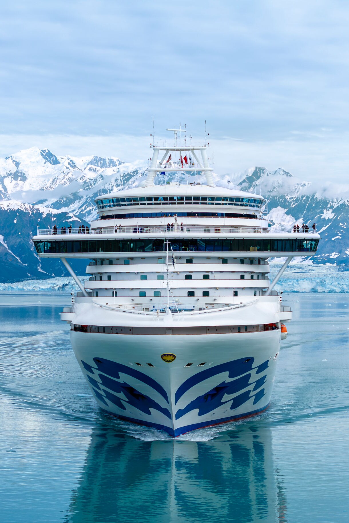 Close-up view of the Crown Princess cruise ship at sea, highlighting its iconic blue wave logo on the bow, with the coastline and white buildings in the background.