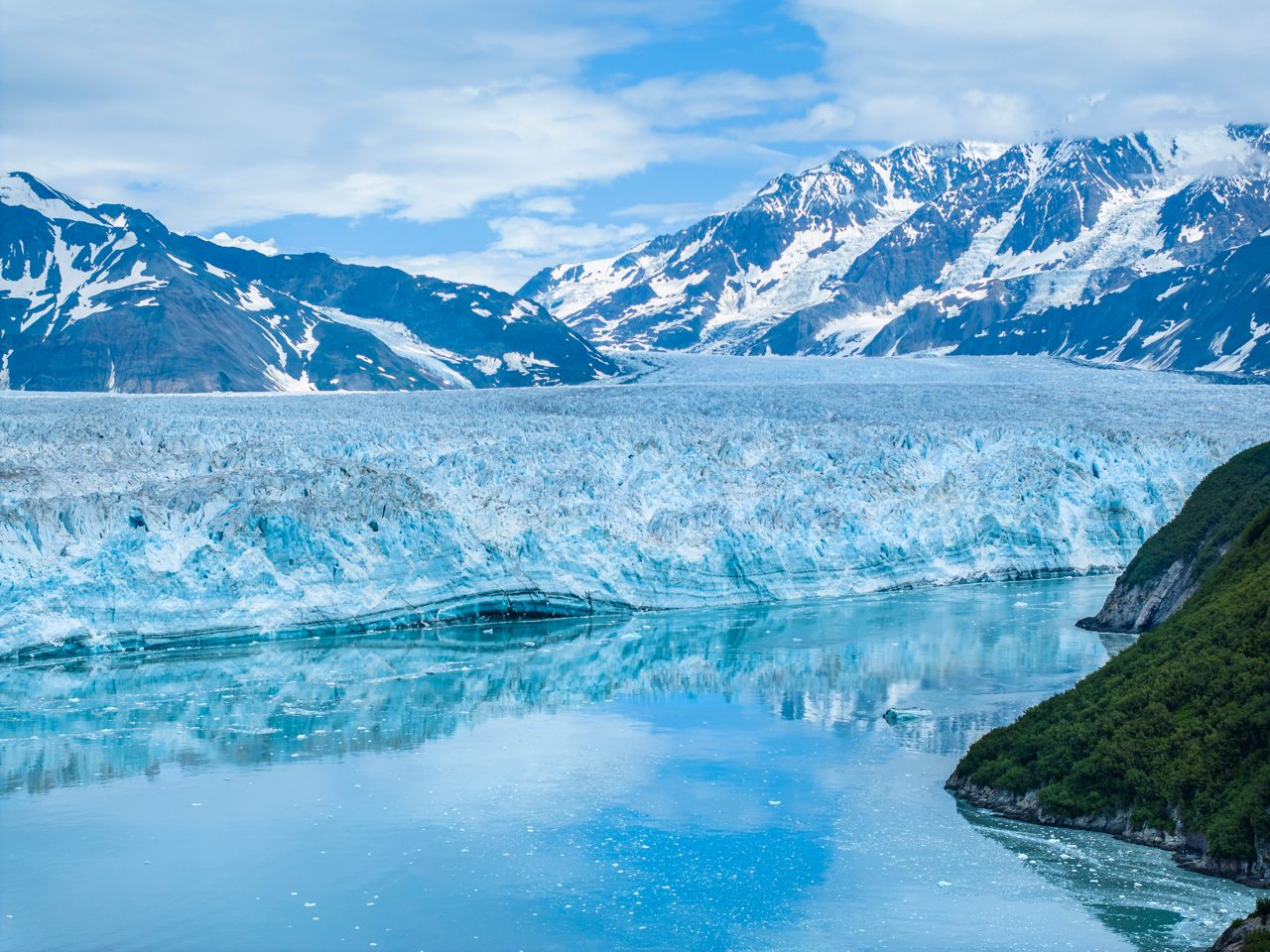The massive turquoise blue face of Hubbard Glacier rises from dark water with a splash of ice and mist near its base, backed by snow-covered mountain peaks.