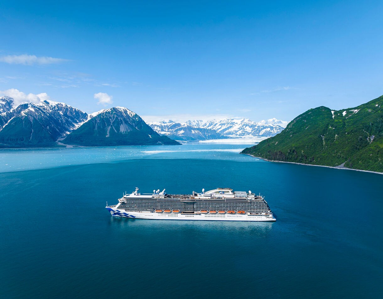A large cruise ship sails through a serene glacier bay surrounded by snow-capped mountains and lush green hills. The calm blue water reflects the stunning natural landscape under a clear sky. The image captures the grandeur of the ship and the pristine beauty of the environment.