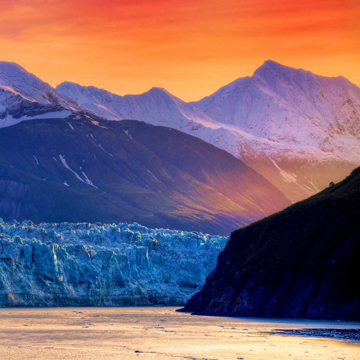 Mountain landscape at golden hour showing snow-capped peaks in warm orange and pink light with a blue glacier in the foreground and calm water reflecting the scene.