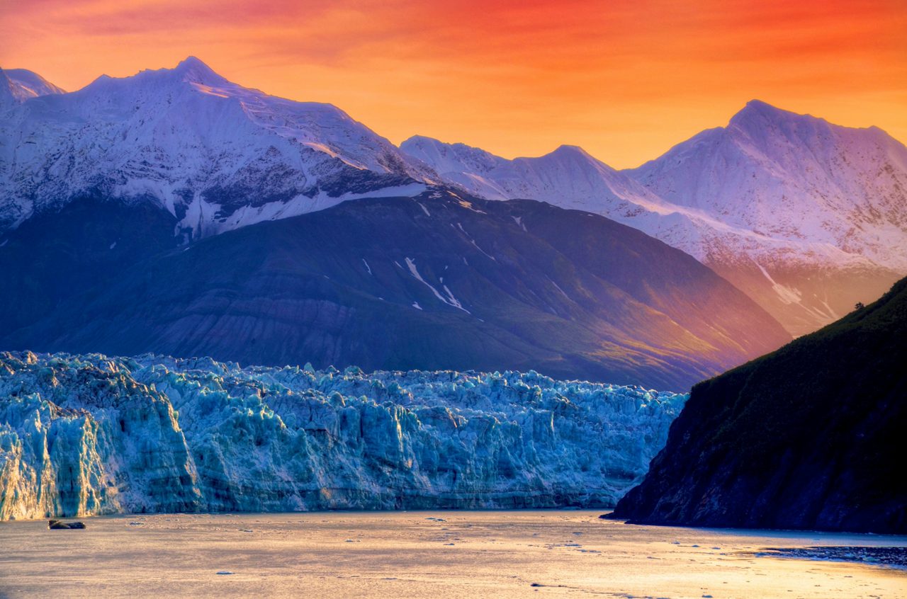 Mountain landscape at golden hour showing snow-capped peaks in warm orange and pink light with a blue glacier in the foreground and calm water reflecting the scene.
