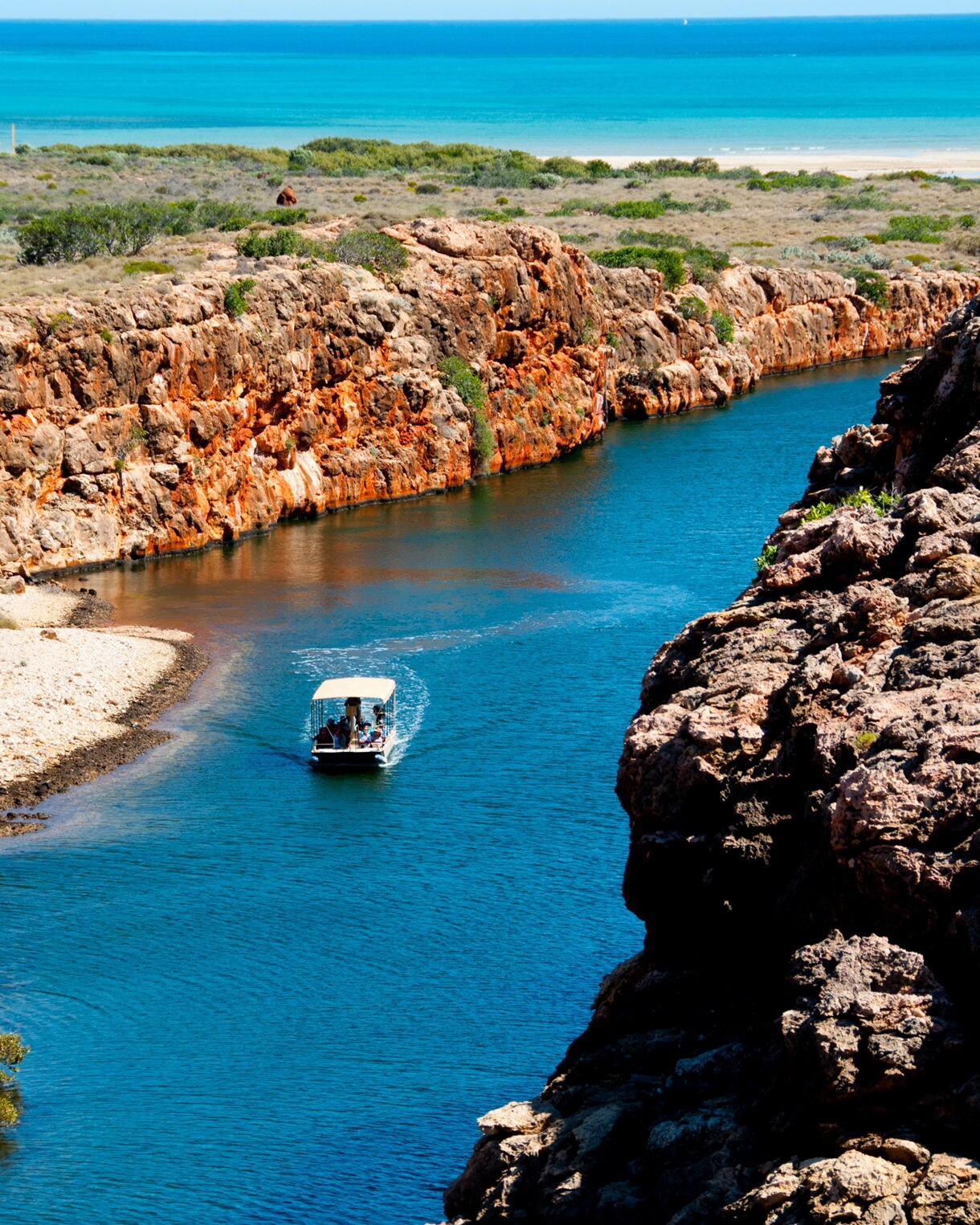 A small boat travels along a deep blue creek bordered by rugged orange cliffs and sparse coastal vegetation near the ocean.