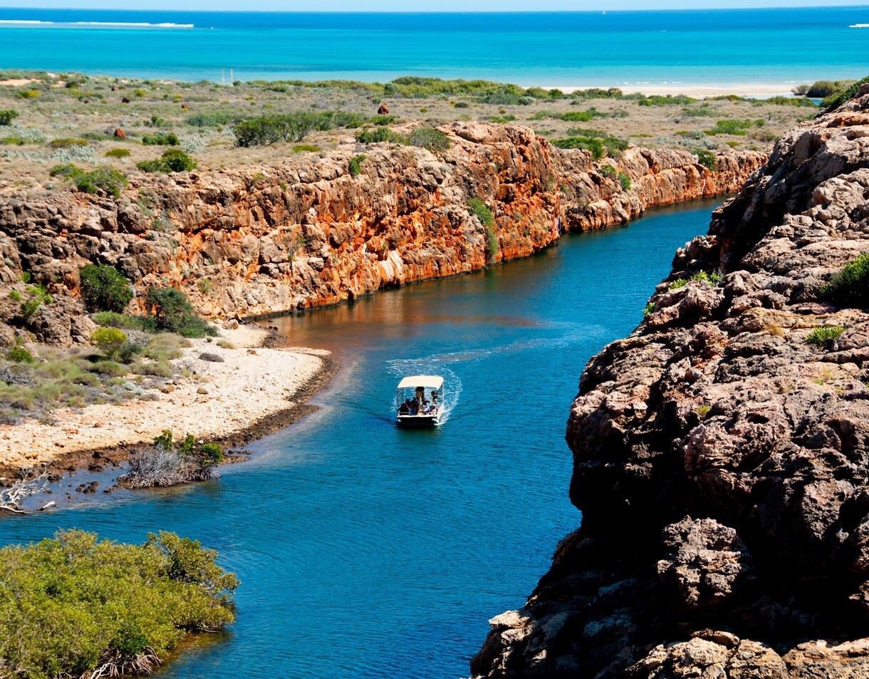 A calm blue creek bordered by rugged orange cliffs and sparse green vegetation under a clear sky.