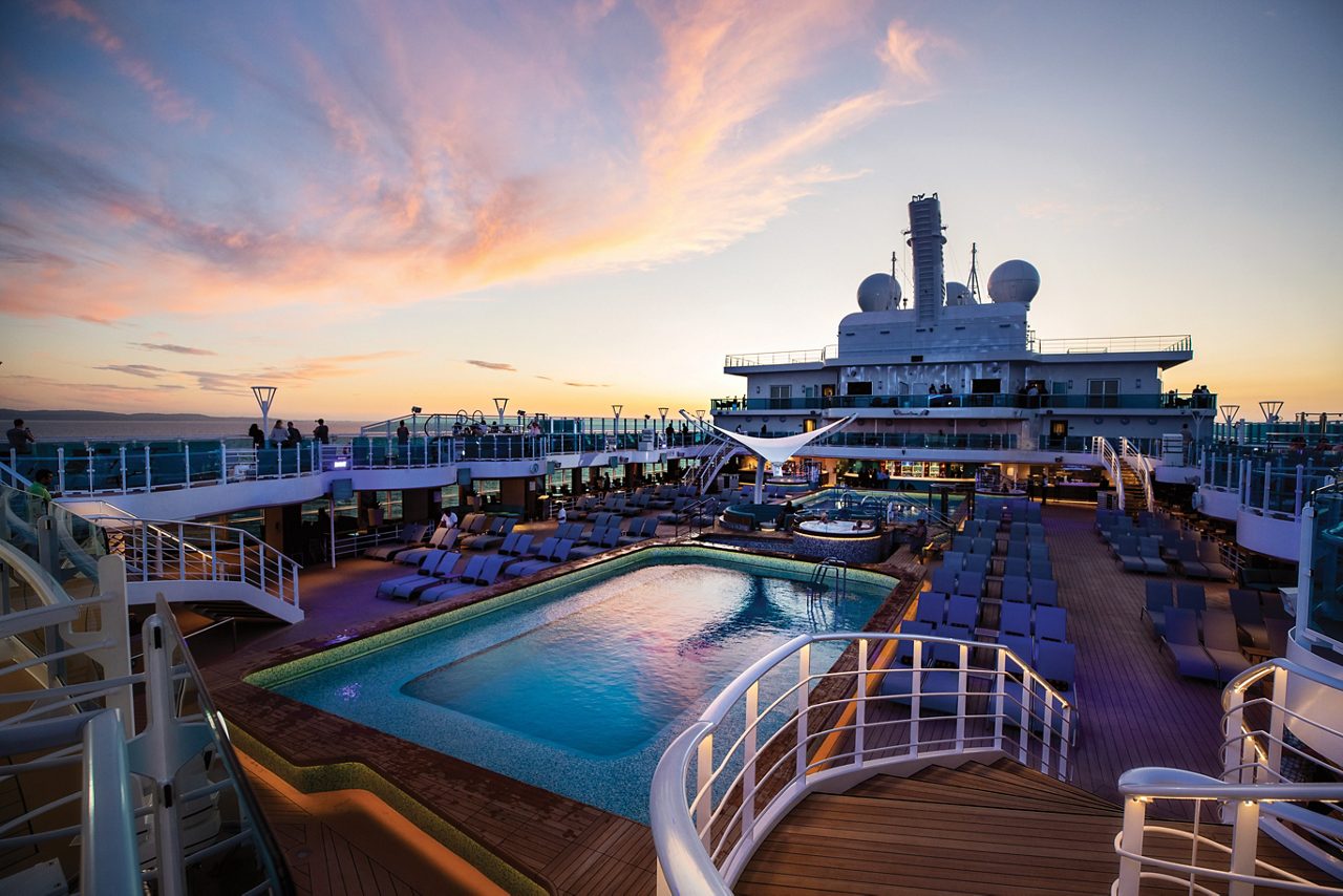  A cruise ship sets sail at sunset, with the ocean reflecting the warm golden hues of the sky. Passengers are seen on the deck enjoying the peaceful view as the ship glides across the water.